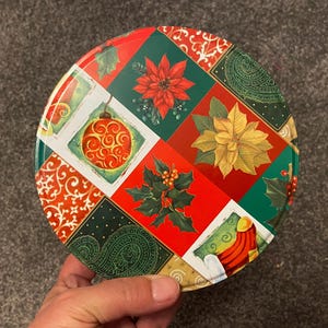 May include: Round Christmas-themed tin with a patchwork design featuring red, green, and gold poinsettias, holly, ornaments, and paisley patterns.