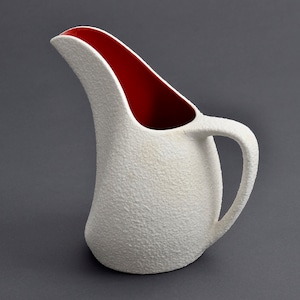 Może przedstawiać: Biały dzbanek ceramiczny z fakturowaną powierzchnią i czerwonym wnętrzem. Dzbanek ma zakrzywiony dzióbek i uchwyt.