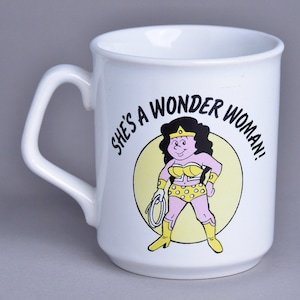 Könnte beinhalten: Weiße Keramiktasse mit einem schwarz-gelben Comicbild von Wonder Woman. Die Tasse trägt den Text "She's a Wonder Woman!" aufgedruckt.
