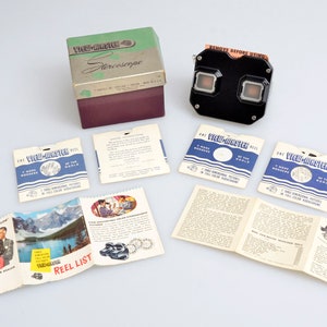 May include: A vintage View-Master stereoscope viewer in black with a red label that says "Remove Before Using". The viewer is surrounded by various View-Master reels, instruction manuals, and a box with the words "View-Master Stereoscope" printed on it.