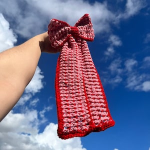 May include: A red and pink crocheted bow with a textured pattern. The bow is held up against a blue sky with white clouds.