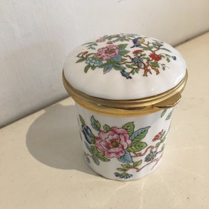 May include: A white porcelain trinket box with a floral design and gold trim. The lid has a hinged design and features a floral pattern with a small bird.