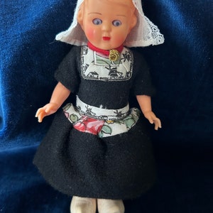 Puede incluir: Una muñeca holandesa vintage que lleva un vestido tradicional blanco y negro con un delantal de estampado floral. La muñeca tiene un gorro de encaje blanco y zapatos de madera.