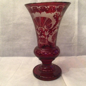 Può includere: Un vaso di vetro rosso rubino con la parte superiore svasata e base rotonda. Il vaso presenta intricati disegni bianchi incisi, tra cui motivi floreali e architettonici. Il vaso ha una sezione centrale bulbosa e un bordo smerlato.