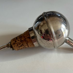 Peut inclure: Un bouchon de bouteille en argent avec un insert en liège et un dessus rond et bulbeux. Le dessus est réfléchissant et montre une réflexion floue d'une pièce.