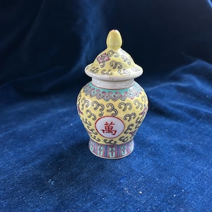 Pode incluir: Um pequeno pote de cerâmica amarelo com tampa. O pote é decorado com um padrão repetitivo de redemoinhos e um caractere chinês vermelho. A tampa tem um pequeno botão e desenhos florais. A base tem um padrão listrado colorido.