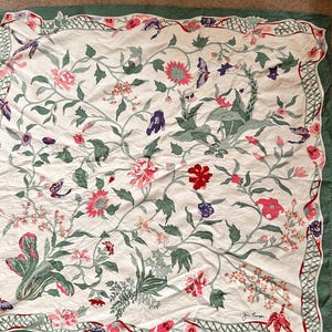 Puede incluir: Textil crema con un estampado floral que presenta flores rosas, rojas y moradas, hojas verdes y mariposas. El dise&ntilde;o est&aacute; enmarcado por un borde verde con un borde festoneado. Es probable que el textil sea un art&iacute;culo decorativo, como una bufanda o un mantel.
