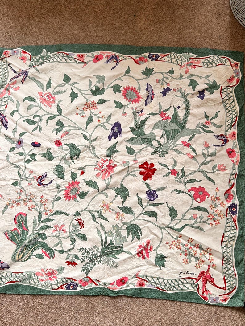 Puede incluir: Textil crema con un estampado floral que presenta flores rosas, rojas y moradas, hojas verdes y mariposas. El dise&ntilde;o est&aacute; enmarcado por un borde verde con un borde festoneado. Es probable que el textil sea un art&iacute;culo decorativo, como una bufanda o un mantel.