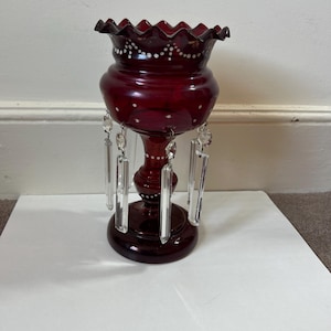 Puede incluir: Un jarrón de cristal rojo rubí con borde ondulado y prismas de cristal colgantes. El jarrón tiene un cuerpo bulboso y una base de pedestal, con detalles de puntos blancos. Una pieza decorativa.