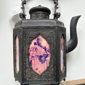 Puede incluir: Una tetera de metal negro con tapa abatible y asa. La tetera tiene un cuerpo cuadrado con paneles decorativos que presentan escenas pintadas de personas con vestimenta tradicional china.