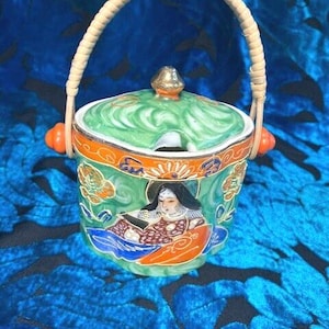 Puede incluir: Un azucarero de cerámica vintage con tapa y asa trenzada. El cuenco está decorado con un diseño floral verde y naranja y una imagen pintada de una mujer. El asa es de material trenzado.