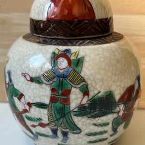 Könnte beinhalten: Eine weiße Keramikdose mit Deckel, verziert mit einer Craquelé-Glasur und handbemalten Figuren in Rot, Grün, Blau und Gelb. Die Figuren tragen traditionelle chinesische Kleidung und sind in einer Landschaft dargestellt.