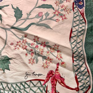 Puede incluir: Un pa&ntilde;uelo de seda con un dise&ntilde;o floral con flores rosas y rojas, hojas verdes y un p&aacute;jaro rojo. El pa&ntilde;uelo tiene un fondo blanco con un borde verde y la firma "Jim Thompson".