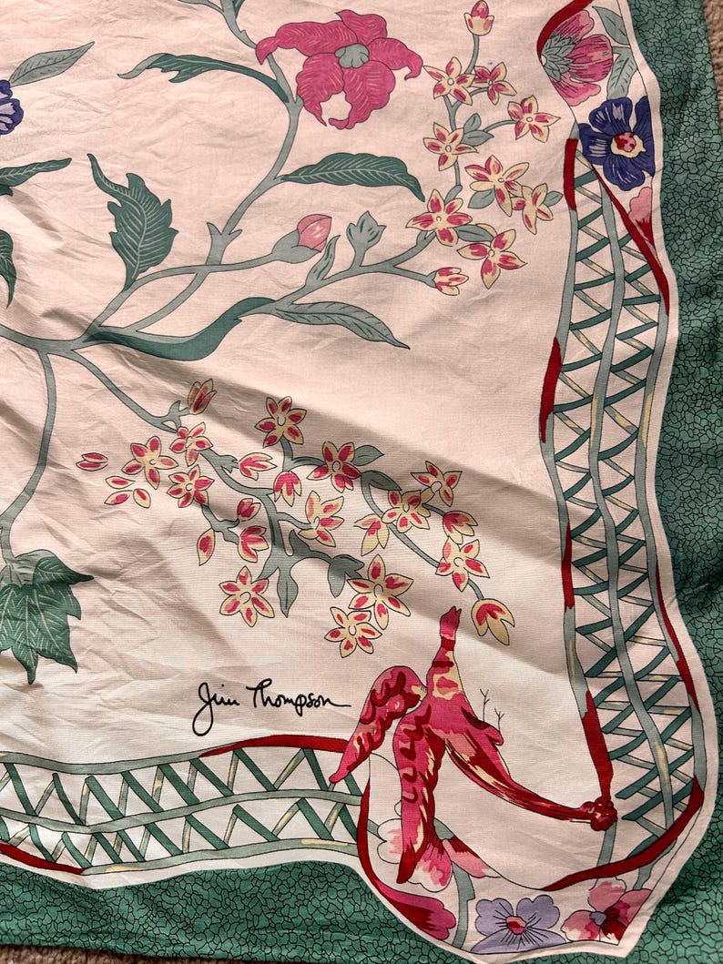 Puede incluir: Un pa&ntilde;uelo de seda con un dise&ntilde;o floral con flores rosas y rojas, hojas verdes y un p&aacute;jaro rojo. El pa&ntilde;uelo tiene un fondo blanco con un borde verde y la firma "Jim Thompson".
