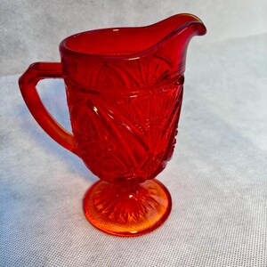 May include: A red glass creamer with a footed base and a cut glass design.