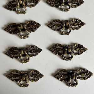 May include: Eight antique brass drawer pulls with ornate, filigree-style designs. Each pull features a decorative backplate and a ring-shaped handle. The hardware has a vintage aesthetic.