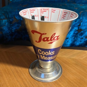 May include: A gold and blue Tala Cooks Dry Measure cup with a silver base. The cup has a graduated scale for measuring ingredients like desiccated coconut, cornflour, cocoa, almonds, suet, sugar, rice, lentils, barley, and sultanas.