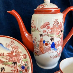 May include: A white teapot with a red lid and handle, decorated with a Japanese-inspired design of women in traditional clothing and a landscape. The teapot is part of a tea set that includes two cups and saucers.