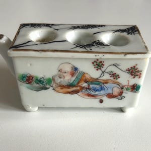 Puede incluir: Tintero rectangular de cerámica blanca con tapa con dos aberturas circulares. El tintero está decorado con una colorida ilustración de una figura reclinada y detalles florales. La tapa tiene un borde pintado de negro y el tintero tiene tres patas pequeñas.