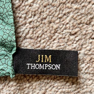 Puede incluir: Una etiqueta de tela negra con el texto dorado "JIM THOMPSON" se muestra sobre un fondo beige texturizado. La etiqueta est&aacute; adherida a una secci&oacute;n de tela con un patr&oacute;n geom&eacute;trico verde y negro.