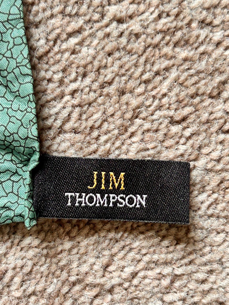 Puede incluir: Una etiqueta de tela negra con el texto dorado "JIM THOMPSON" se muestra sobre un fondo beige texturizado. La etiqueta est&aacute; adherida a una secci&oacute;n de tela con un patr&oacute;n geom&eacute;trico verde y negro.