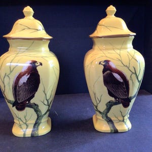 May include: A pair of yellow ceramic urns with lids, each decorated with a painted eagle on a branch. The urns have a gold trim and a branch design. The background is dark blue.