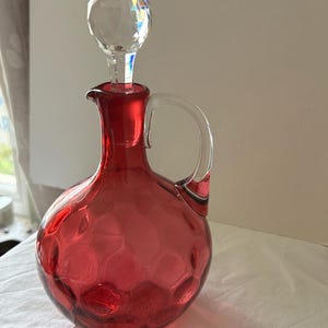 May include: A red glass decanter with a crystal stopper. The decanter has a round body with a textured pattern and a clear glass handle. The stopper is a faceted crystal ball. The decanter is likely used for serving beverages.