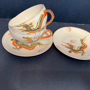 May include: Two white porcelain teacups and two matching saucers featuring hand-painted dragon motifs in shades of orange, black, and gold. The cups have curved handles with orange accents and gold trim along the rims and edges of the saucers.