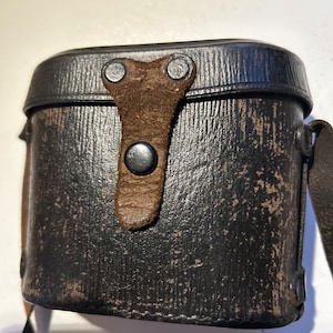 May include: A black leather case with a brown leather strap and metal clasps. The case has a worn and distressed look.