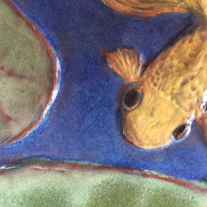 Puede incluir: Un primer plano de una baldosa de cerámica con una imagen pintada de un pez dorado amarillo nadando en agua azul. El pez dorado está mirando al espectador y tiene un ojo negro. La baldosa tiene un fondo verde y rojo.