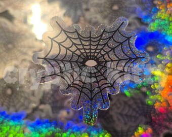 May include: A holographic spiderweb sticker with a black web design and a glittery, iridescent finish. The sticker has a unique, irregular shape and is set against a background of similar stickers and colorful bokeh.