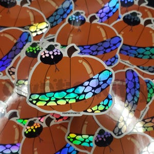 Könnte beinhalten: Orangefarbene Kürbisaufkleber mit schillernden Akzenten. Jeder Aufkleber zeigt einen Kürbis mit einem dekorativen Band und einem kleinen Vogel. Die Aufkleber haben eine glänzende Oberfläche und sind über das Bild verstreut. Der Text "pastelphoenix" ist sichtbar.