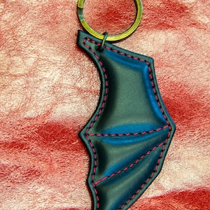 Może przedstawiać: Czarny skórzany brelok w kształcie skrzydła nietoperza z czerwonymi szwami. Brelok ma mosiężny kółko na klucze.