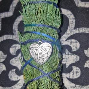 May include: A green bundle of dried herbs tied with blue twine and a silver heart charm. The charm has a Celtic knot design.