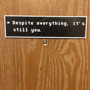May include: A white sign with black text on a wooden door reads "Despite everything, it's still you."
