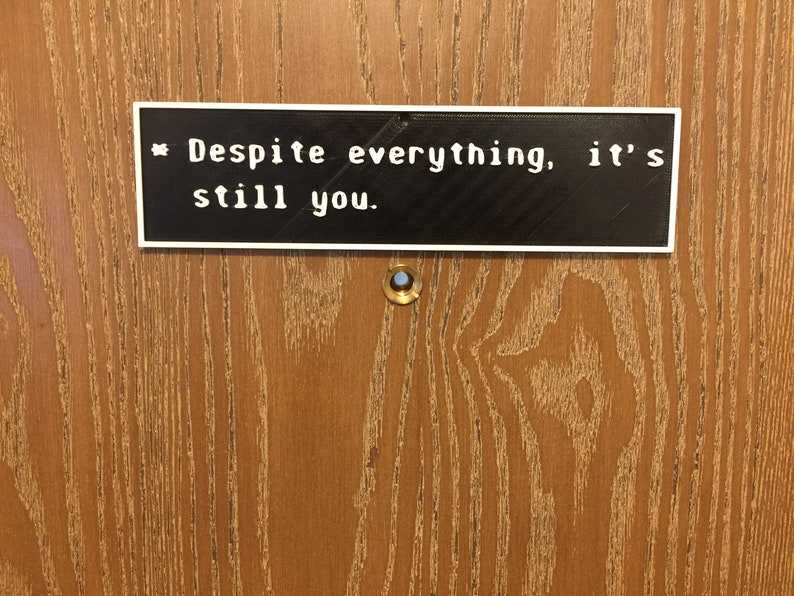May include: A white sign with black text on a wooden door reads "Despite everything, it's still you."