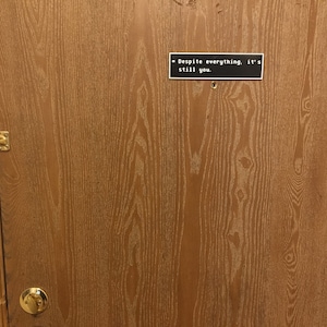 May include: A wooden door with a black sign that reads "Despite everything, it's still you."