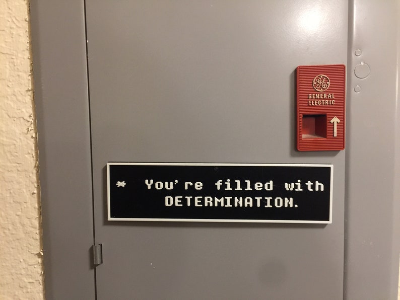 May include: A black and white sign with the text "You're filled with DETERMINATION." on a gray metal door with a red General Electric switch.