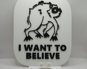 May include: A white rectangular sign with rounded corners featuring a black raised graphic of a creature and the black text I WANT TO BELIEVE. The sign is supported by a white stand with a circular cutout at the base and a small hole at the top.