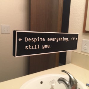 May include: A black and white sign with the text "Despite everything, it's still you." The sign is hanging on a mirror in a bathroom.