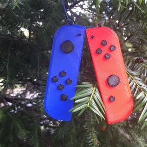 Puede incluir: Un adorno de mando de videojuego azul y rojo, que se asemeja a un Joy-Con, cuelga de una cuerda azul sobre un fondo de follaje verde. Los mandos tienen botones negros y probablemente están impresos en 3D.