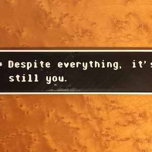 May include: A black rectangular sign with white text that reads "Despite everything, it's still you."