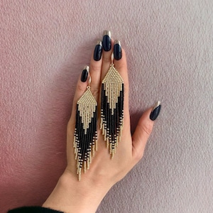 May include: A pair of black and gold beaded earrings with a fringe design. The earrings are hanging from a person's hand.