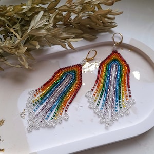 May include: A pair of handmade beaded earrings in a rainbow color scheme. The earrings feature a cascade of colorful beads, with a silver fringe at the bottom. The earrings are displayed on a white and beige oval tray.