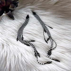 May include: A pair of long, black and white beaded earrings. The earrings are made of small beads that are strung together in a checkered pattern. The earrings are very long and dangle down to the shoulders.