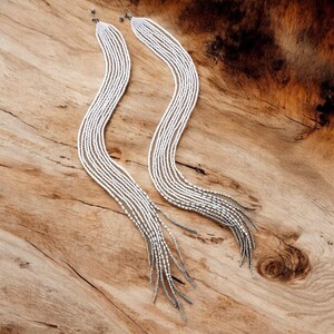 May include: Two long silver beaded necklaces with a fringe of beads at the bottom. The necklaces are laying on a wooden surface.