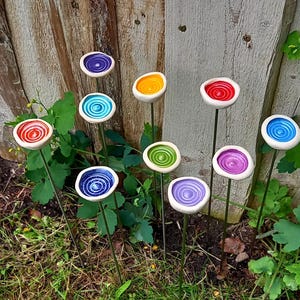 May include: A collection of ten garden stakes featuring circular, bowl-shaped tops in various colors including red, orange, yellow, green, blue, and purple. Each top has a spiral pattern and is mounted on a thin, straight metal rod for display.