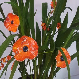 Può includere: Tre fiori di papavero in ceramica arancione con centri neri sono disposti in una pianta in vaso con foglie verdi.