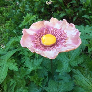 Puede incluir: Una escultura de flor de cerámica con pétalos rosas y morados y un centro amarillo. La flor se encuentra sobre un fondo de follaje verde, creando una exhibición natural y artística. La flor está montada sobre un tallo verde.
