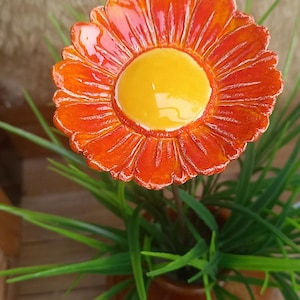 Könnte beinhalten: Eine Keramikblume mit einer leuchtend gelben Mitte, umgeben von strukturierten, glänzenden orangefarbenen Blütenblättern. Die Blume ist auf einem dünnen Stiel befestigt und steht in einem kleinen braunen Topf mit grünen Kunstgrasblättern.